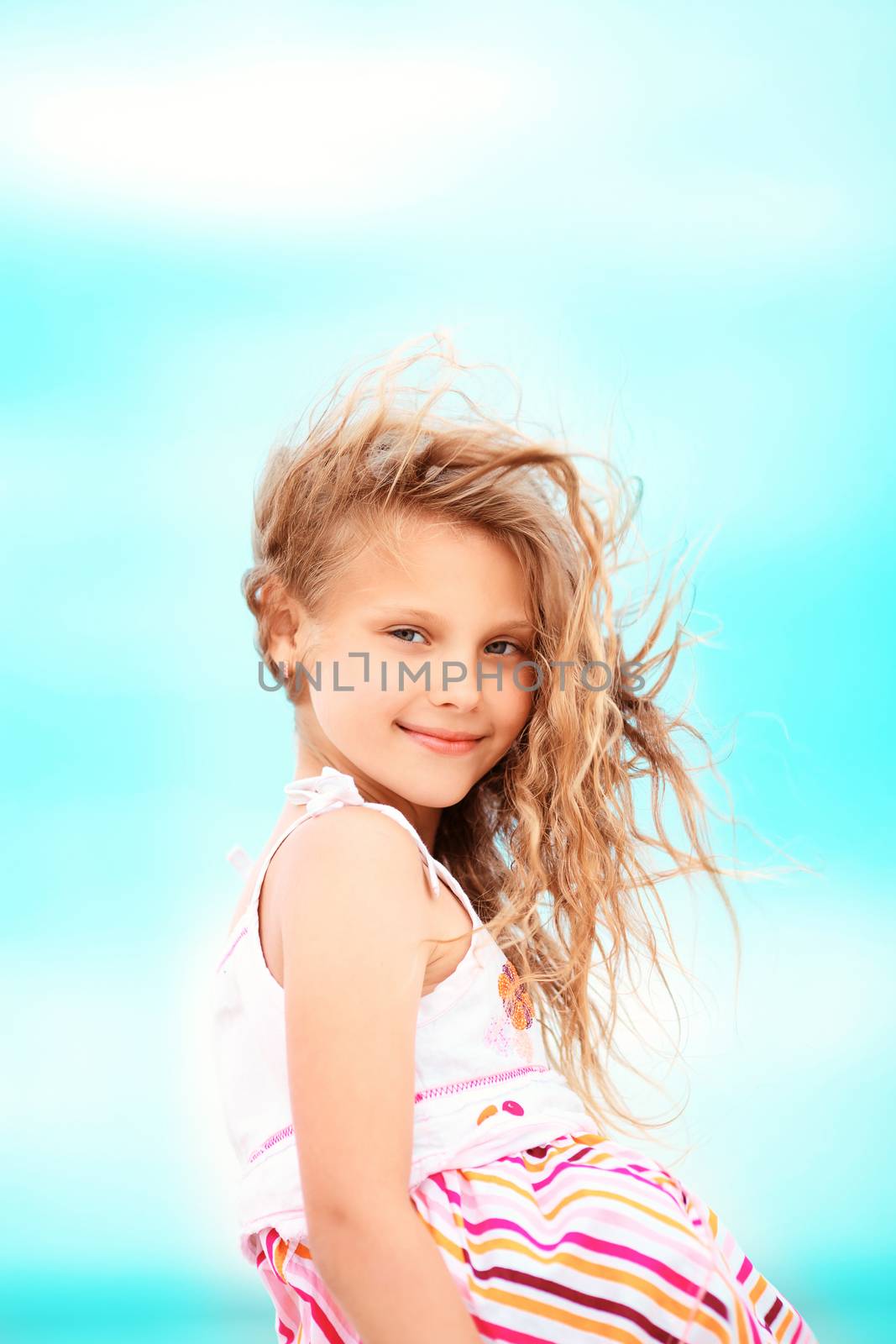 Portrait of a pretty little girl with waving in the wind long hair sitting on the beach against the blue sky