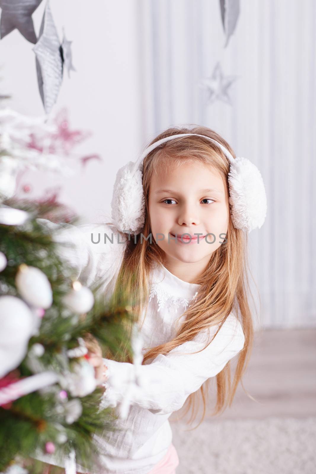 Beautiful little girl in earmuffs decorating Christmas tree with toys and balls. New year preparation.