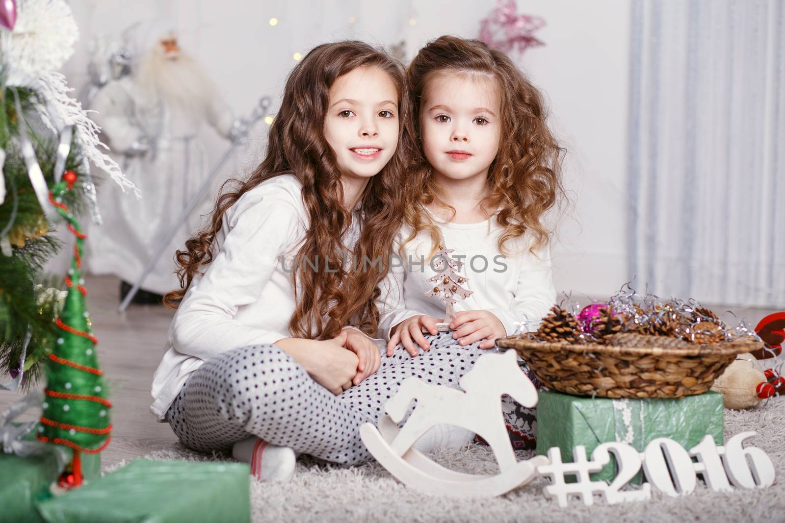 Little girls in comfortable home clothes sitting on floor in beautiful Christmas decorations. Two little sisters decorating Christmas tree with fir-cone. New year preparation. Happy girls and family.