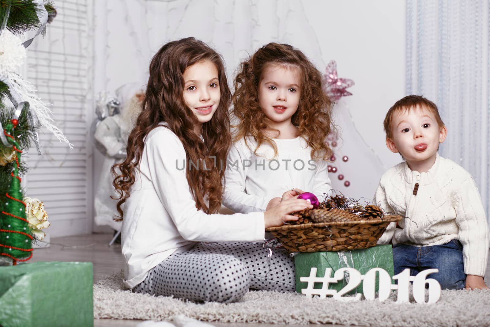 three Little kids in comfortable home clothes sitting on floor in beautiful Christmas decorations. three little siblings decorating Christmas tree with fir-cone. New year preparation. Happy girls and family.