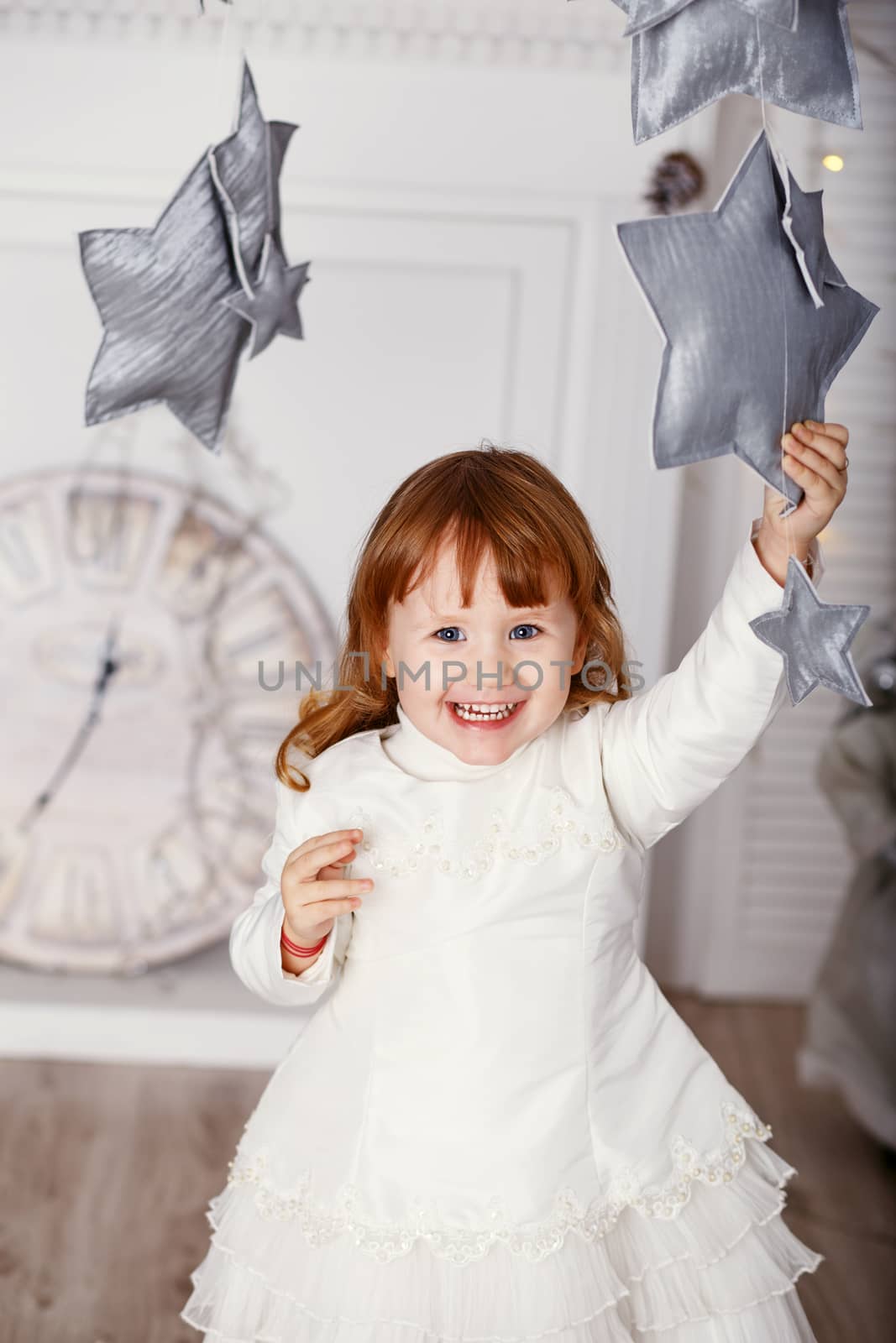 Portrait of a beautiful little girl in a white dress in the interior with Christmas decorations. Baby girl grabs and catches toy star - an element of a Christmas decor