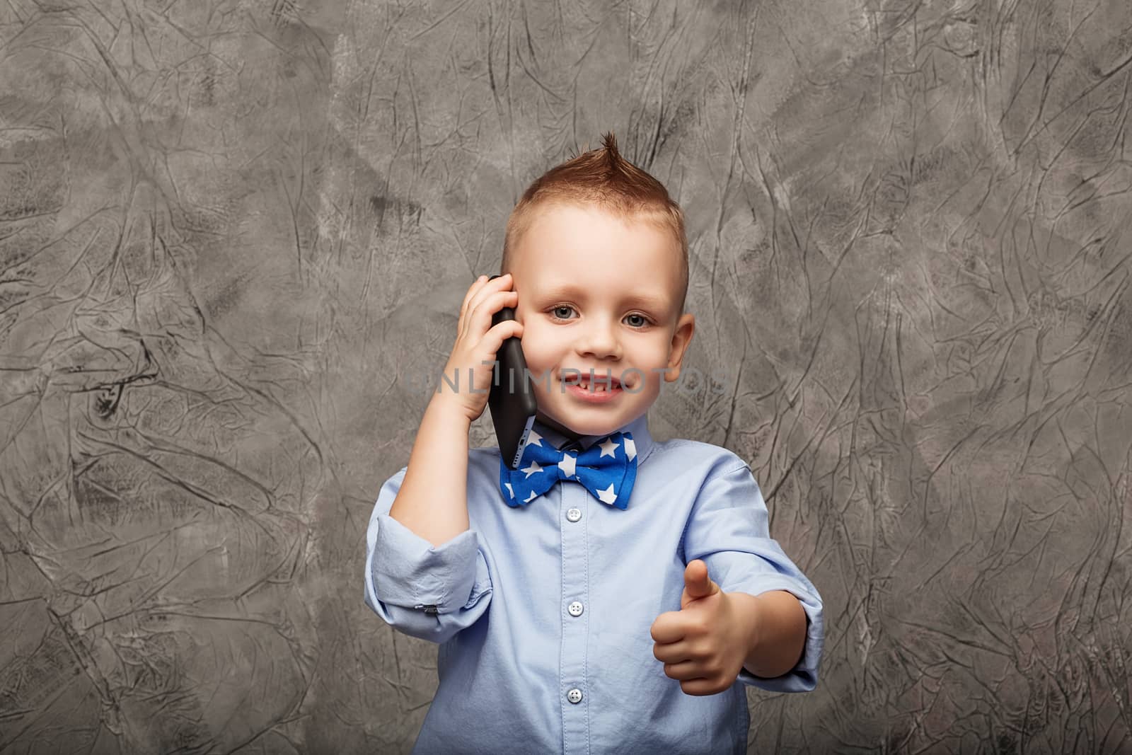Portrait of a cute little boy in blue shirt and bow tie with mobile phone against gray textural background in studio. Kid shows thumb
