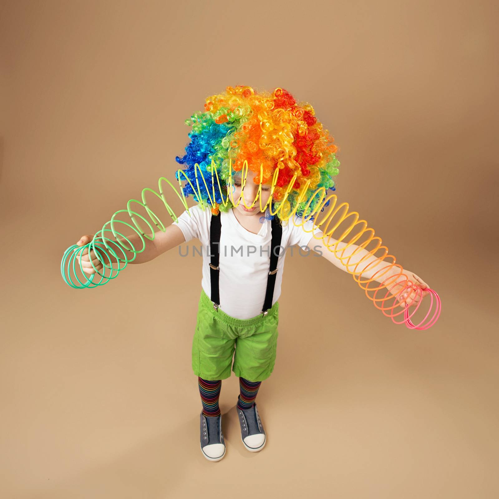 Happy clown boy with large colorful wig. Little boy in clown wig playing with a spring. Portrait of a child shot on a wide-angle lens. Birthday boy. Positive emotions. Top view portrait