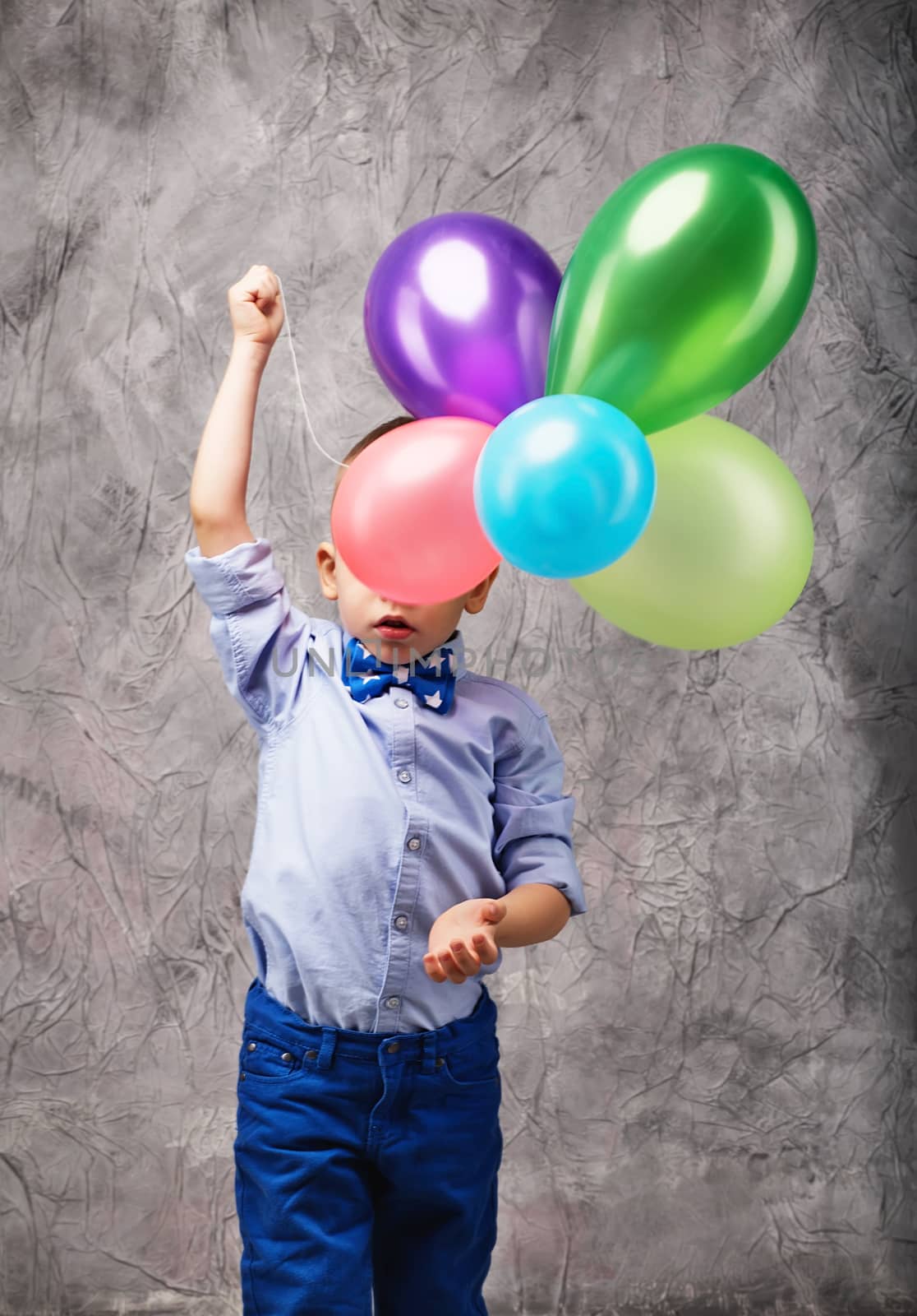 Portrait of a cute little boy in jeans, blue shirt and bow tie with multi-colored balloons in hands on a gray textural background in studio. Birthday boy