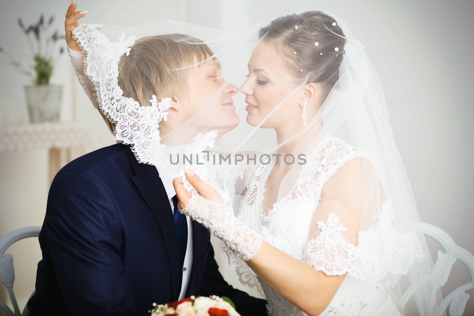 Bride And Groom Kissing Under Veil Holding Flower Bouquet In Hand.