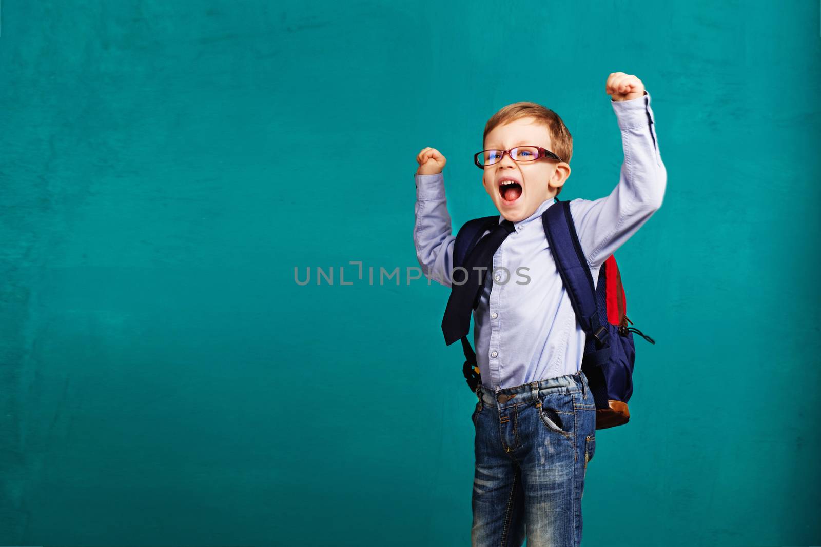 Adorable smiling little kid with big backpack jumping and having fun against blue wall. Looking at camera. School concept. Back to School