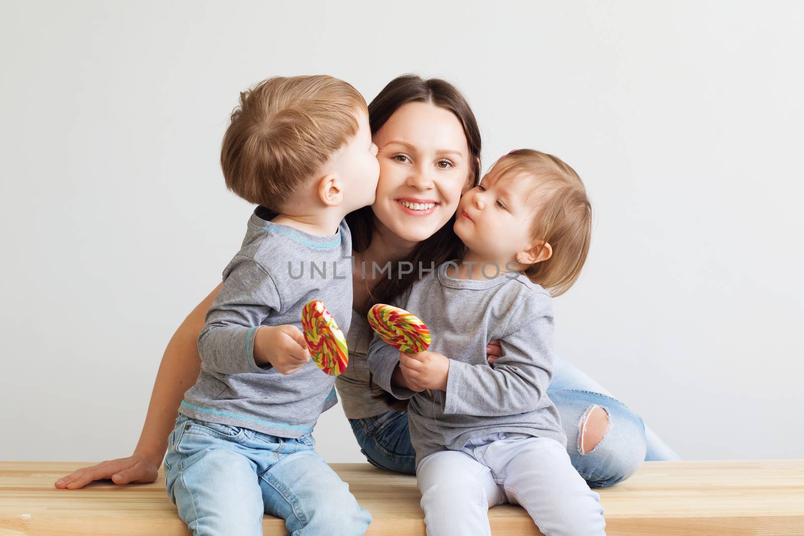 Portrait of a happy mother and her two little children - boy and girl. Happy family against a white background. Little kids kissing mother. Children with lollipops.