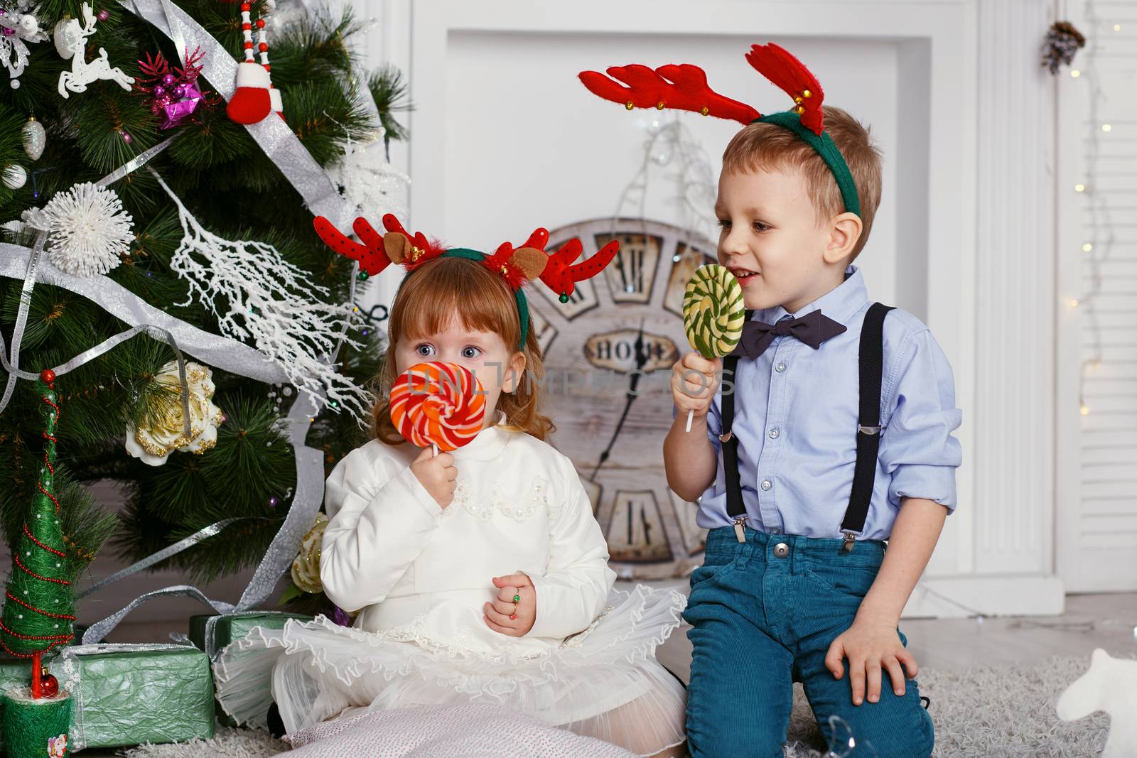 Little kids in reindeer antlers eating a lollipops. Portrait of two funny little kids with delicious candies in the hands. Happy children and family In anticipation of new year and Christmas.