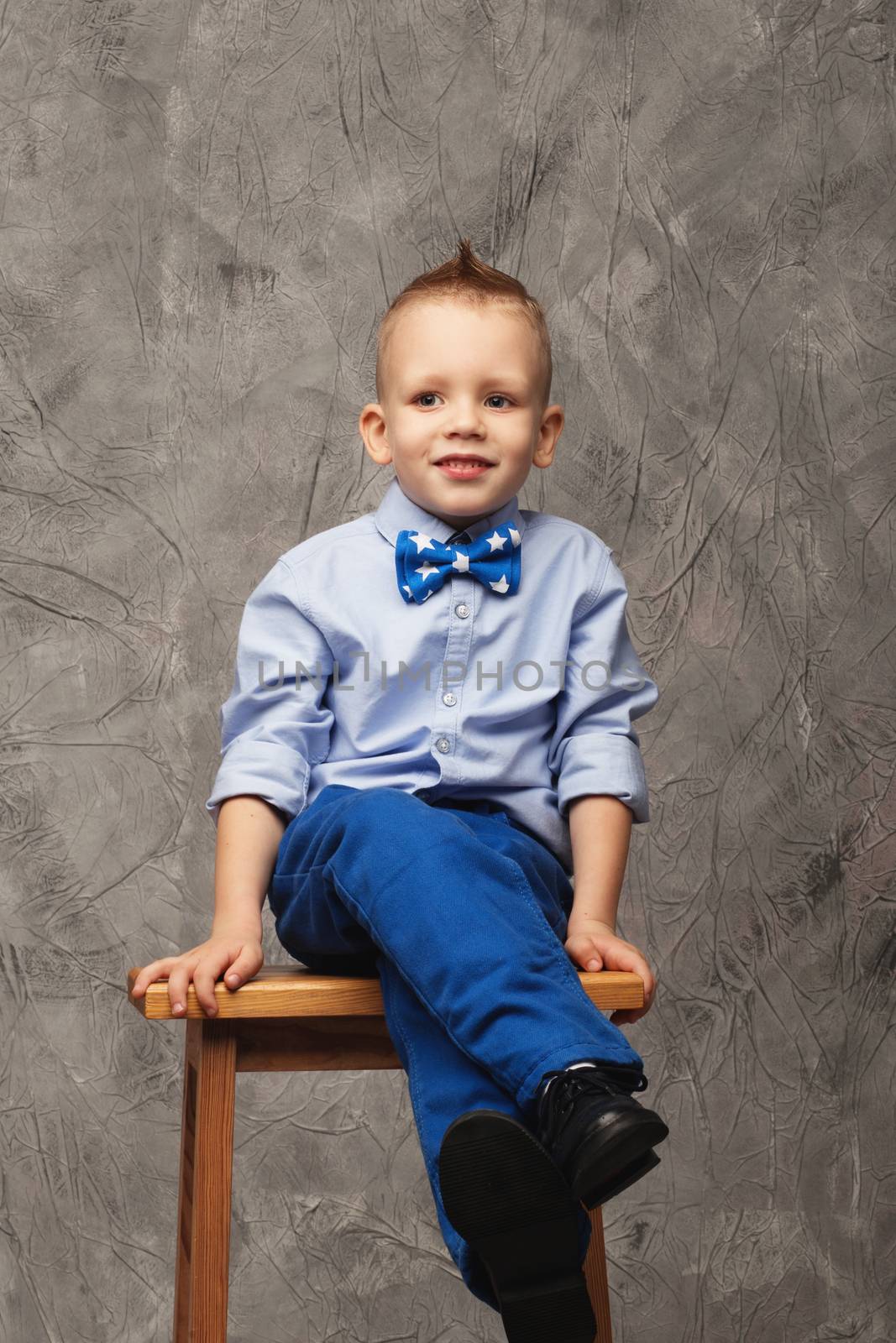 Portrait of a cute little boy in jeans, blue shirt and bow tie on a gray textural background in studio. Portrait of a funny little boy sitting on a high stool