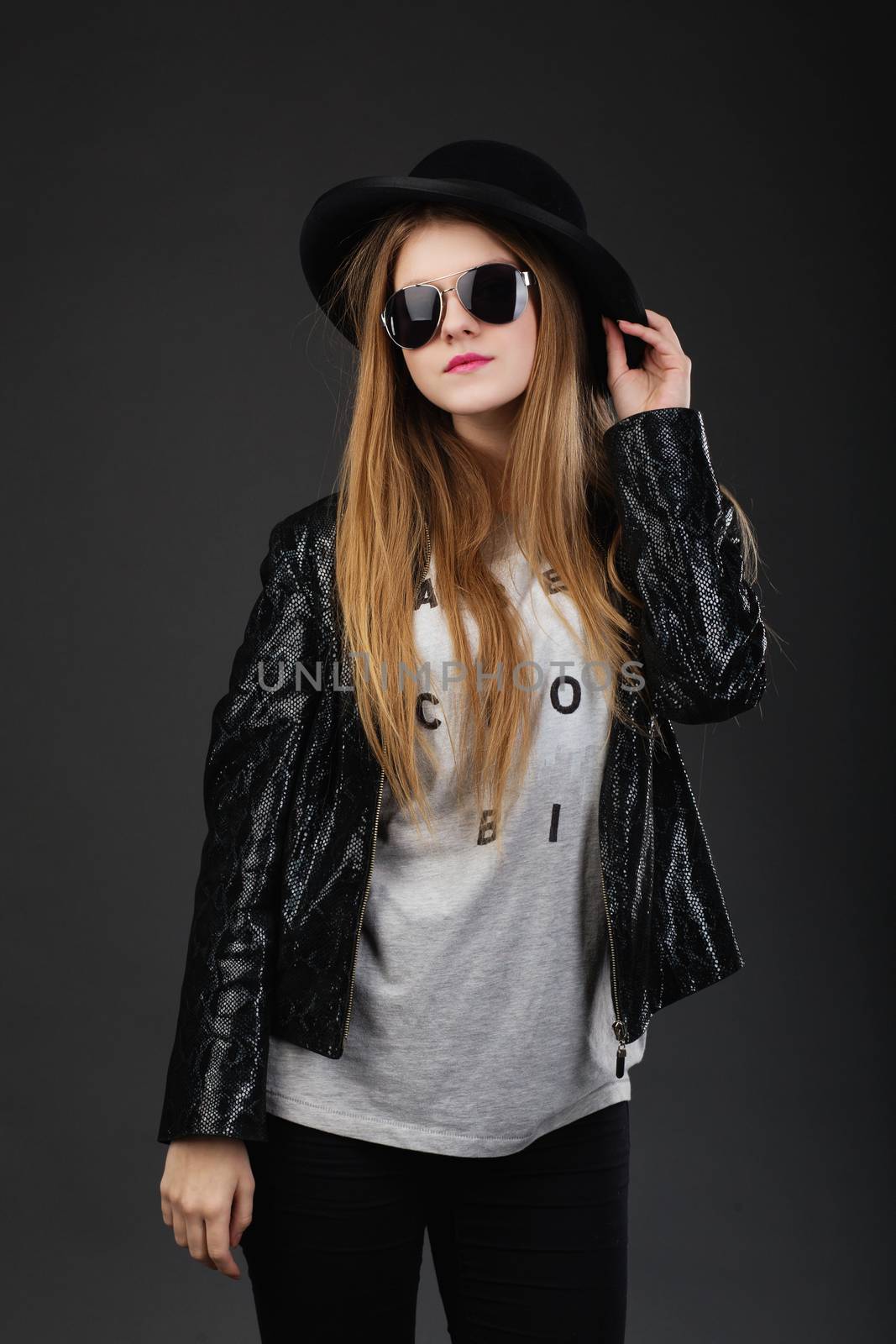 Portrait of beautiful young girl wearing black felt hat, Sunglasses and leather jacket in front of a dark gray background
