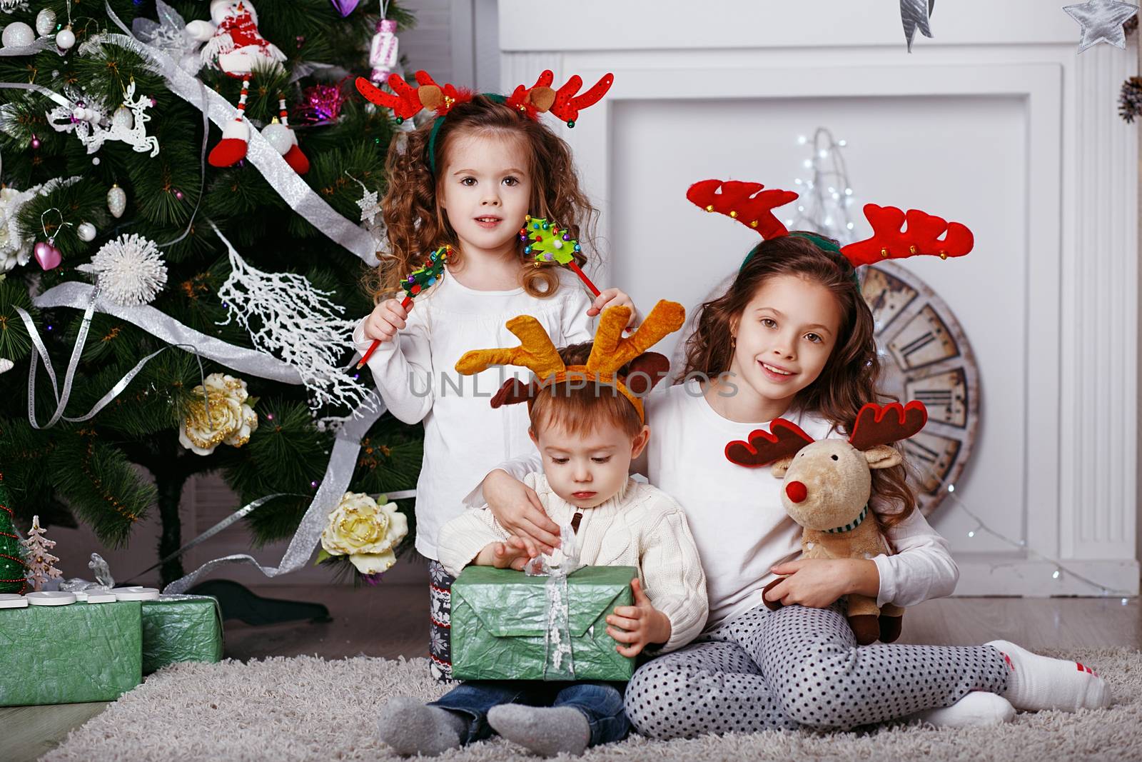Little children in reindeer antlers In anticipation of new year and Christmas. Three little siblings opening christmas gifts. Kids in comfortable home clothes sitting on floor over Christmas tree