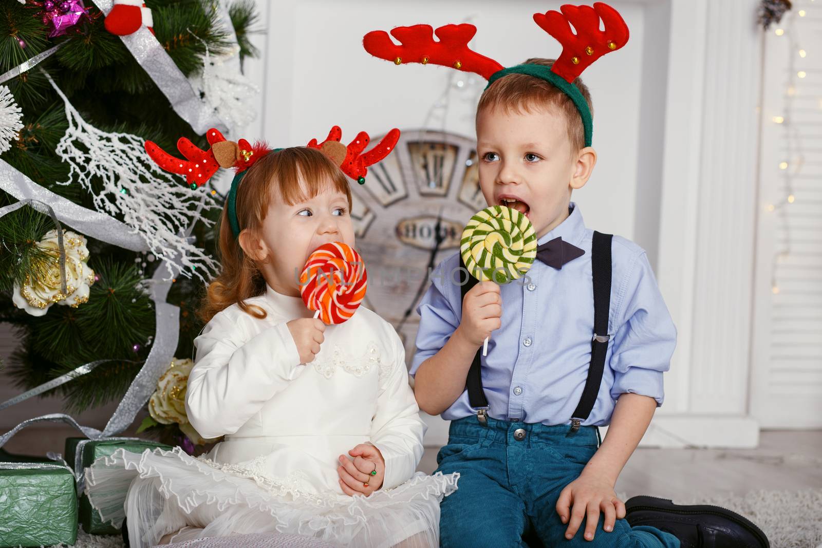 Little girl and boy in reindeer antlers eating a lollipops. Portrait of two funny little kids with delicious candies in the hands. Happy children and family In anticipation of new year and Christmas.