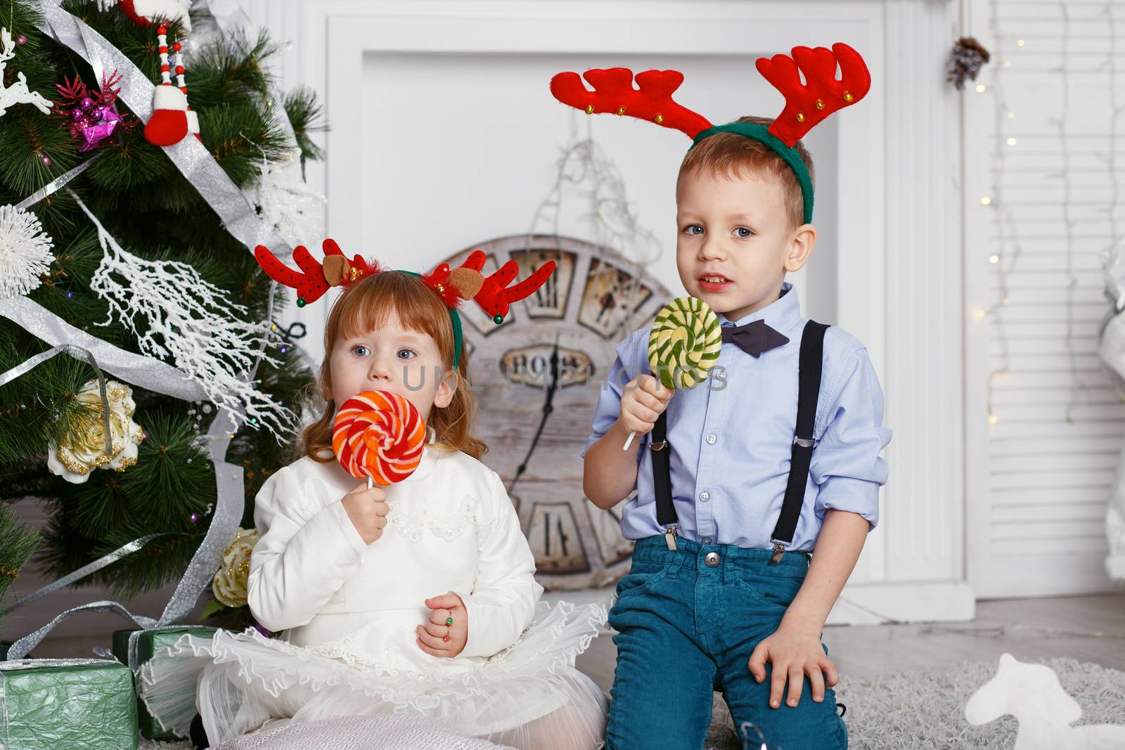 Little children in reindeer antlers eating a lollipops. Portrait of two funny little kids with delicious candies in the hands. Happy children and family In anticipation of new year and Christmas.