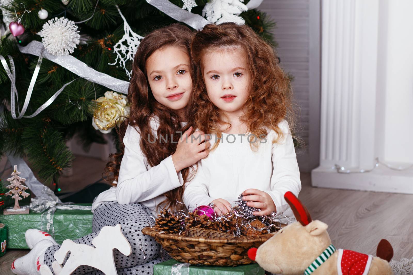 Little girls in comfortable home clothes sitting on floor in beautiful Christmas decorations. Two little sisters decorating Christmas tree with fir-cone. New year preparation. Happy girls and family.