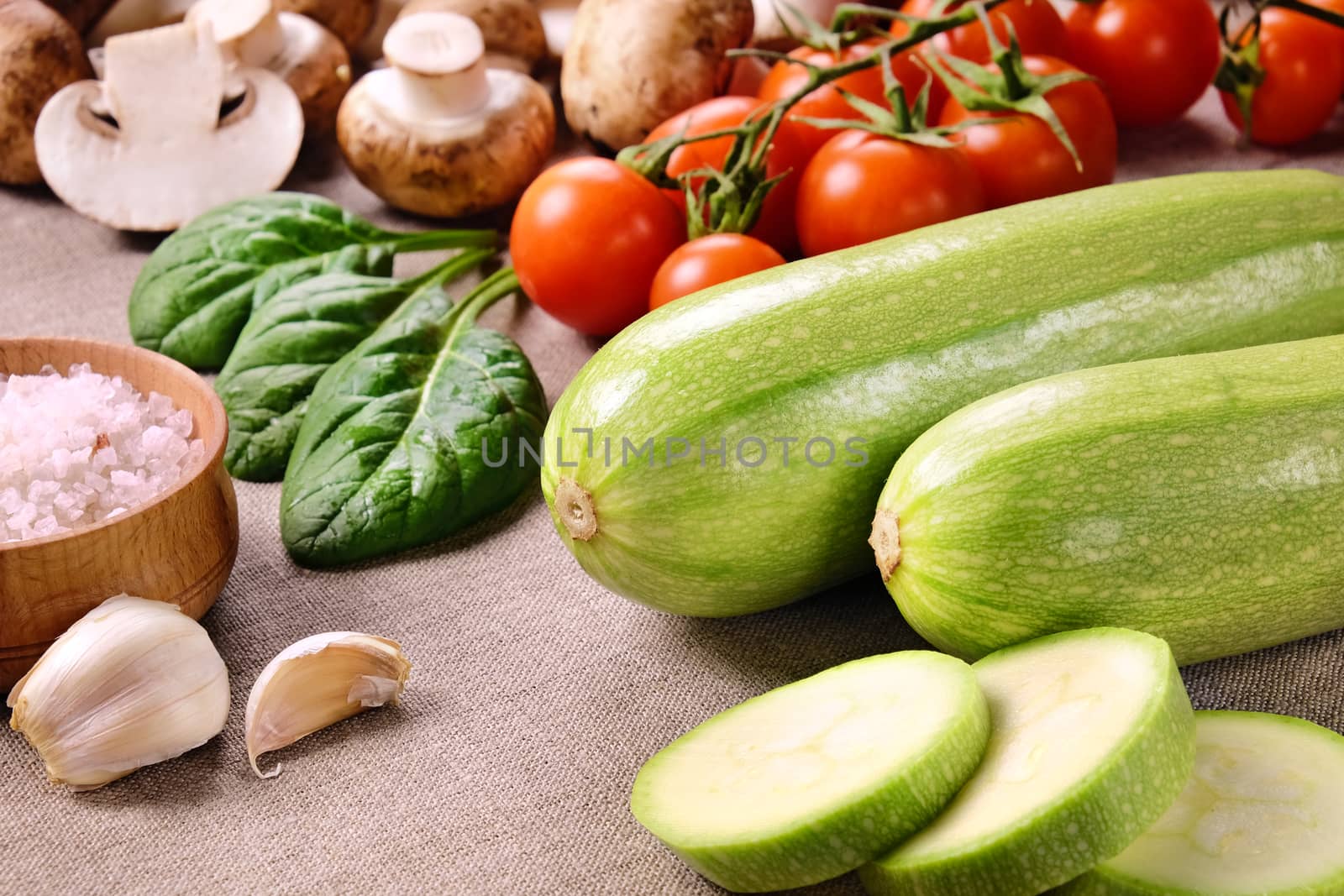 Two whole zucchini and a few sliced pieces, cherry tomatoes, mushrooms, salt in wooden salt shaker, cloves of garlic and spinach leaves on a linen cloth.