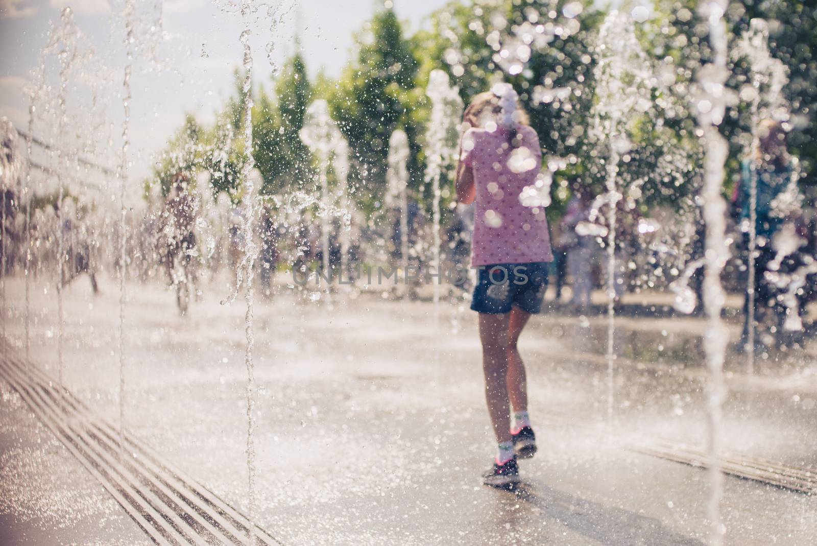 Little cute girl walking in open street fountain at hot sunny day. Back view