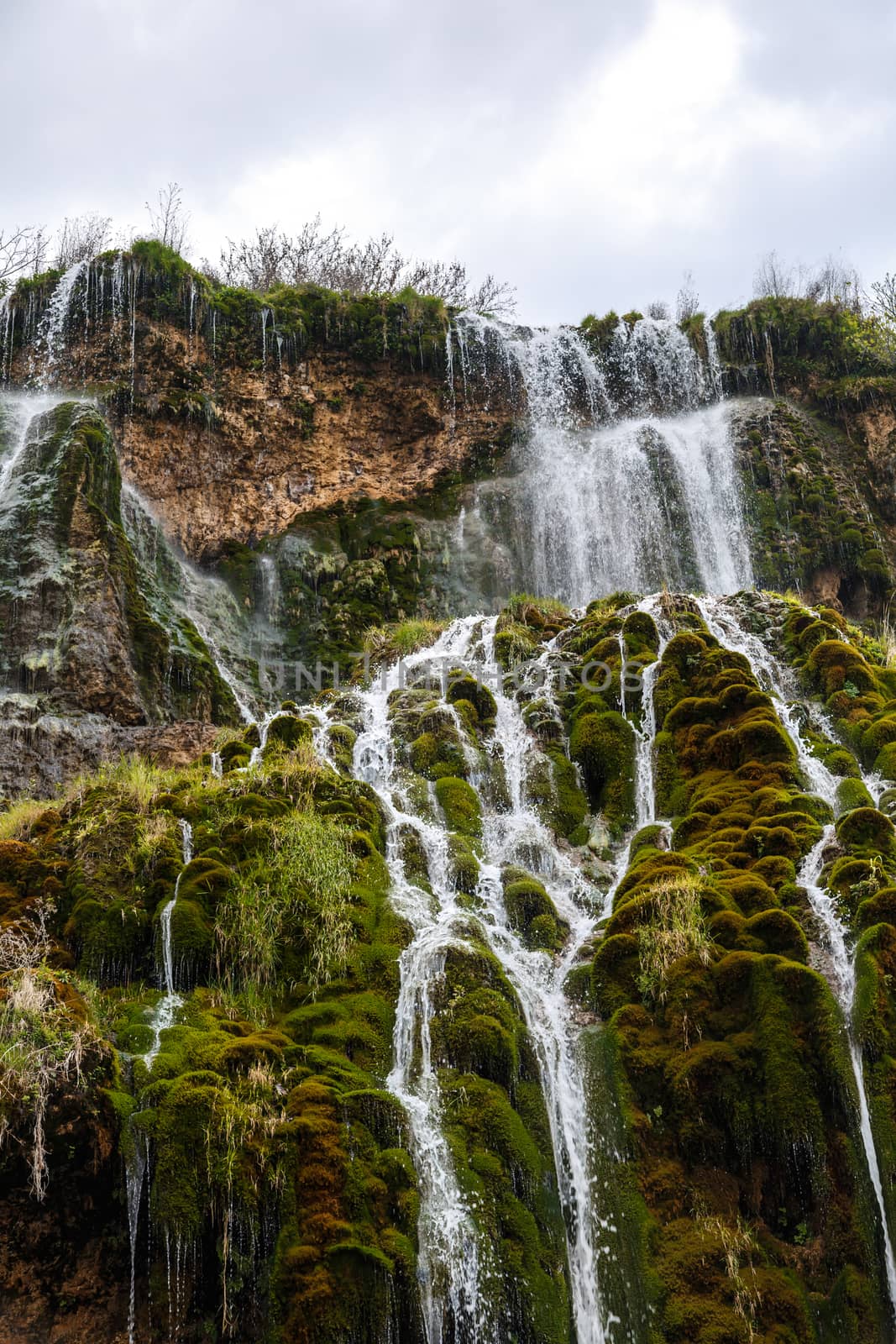 View of a waterfall, flowing from high inside meadow area, with green trees and plants around.