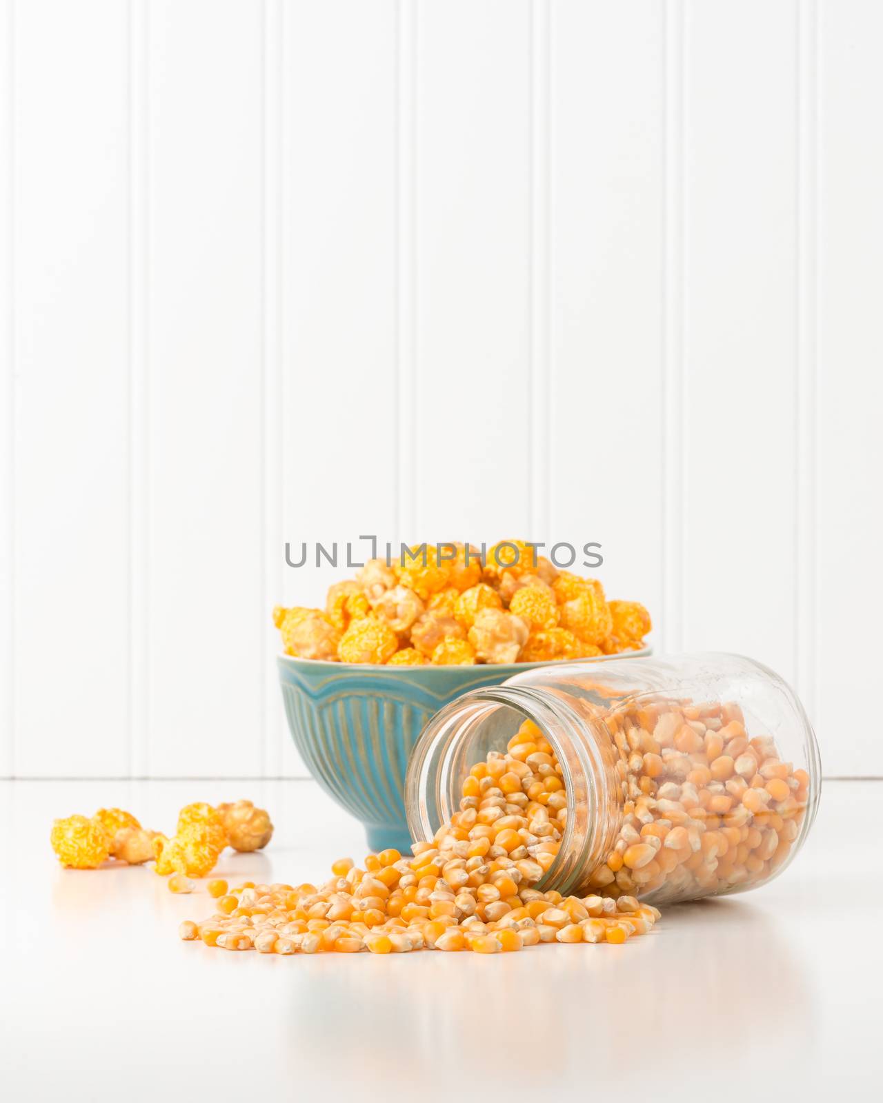 Jar of raw popcorn kernels with a bowl of popped corn in the background.