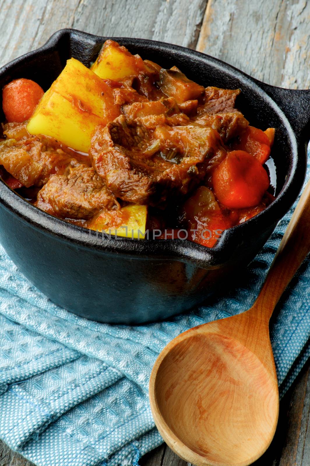 Homemade Beef Stew with Carrots, Potatoes, Celery and Leek in Cast Iron closeup on Blue Napkin with Wooden Spoon 
