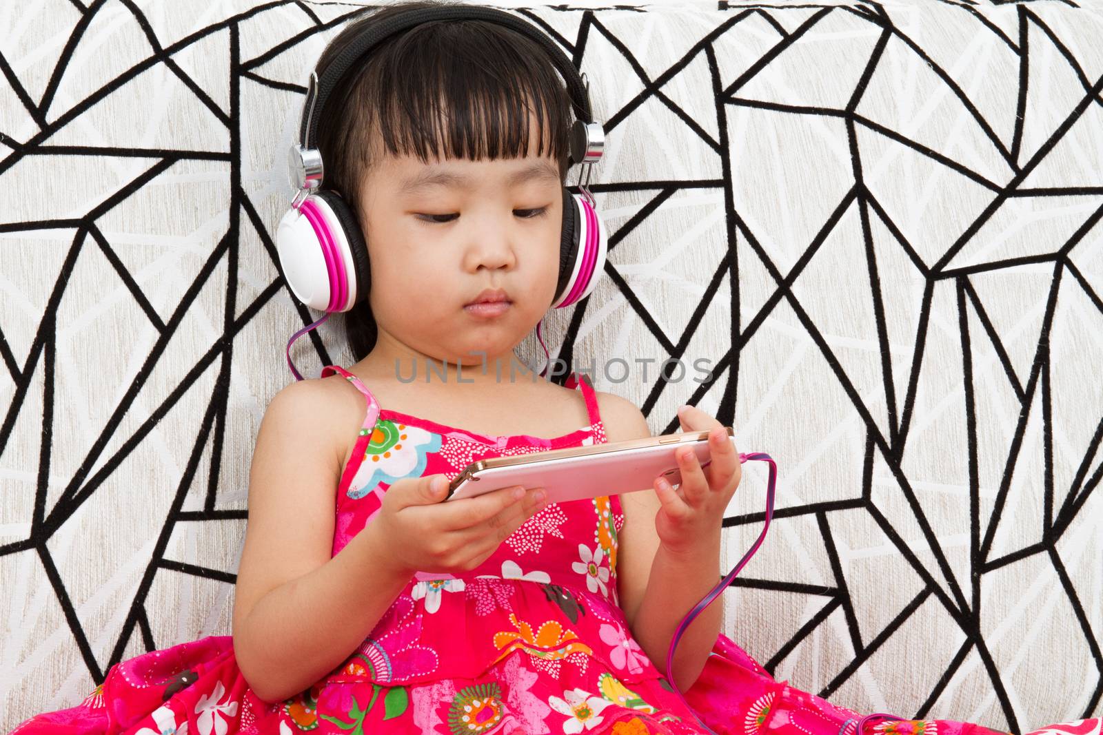 Chinese little girl on headphones holding mobile phone sitting on sofa