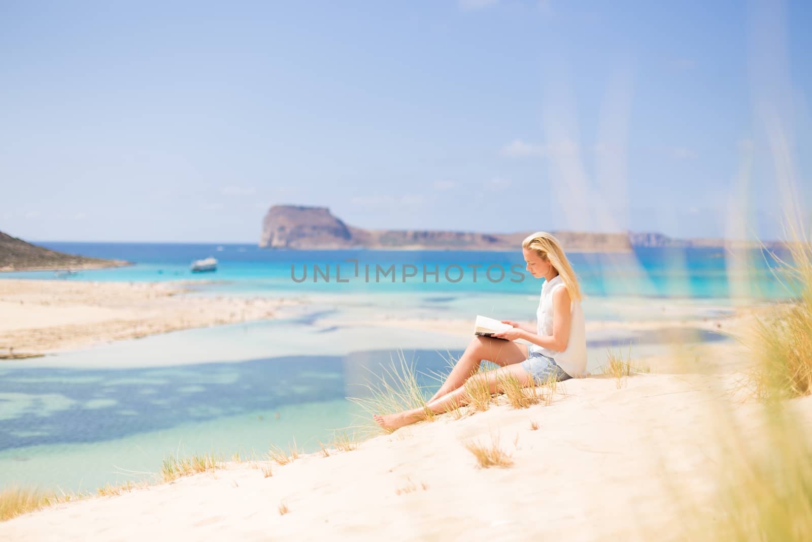 Relaxed woman enjoying sun, freedom and good book an beautiful sandy beach of Balos in Greece. Young lady reading, feeling free and relaxed. Vacations, freedom, happiness, enjoyment and well being.