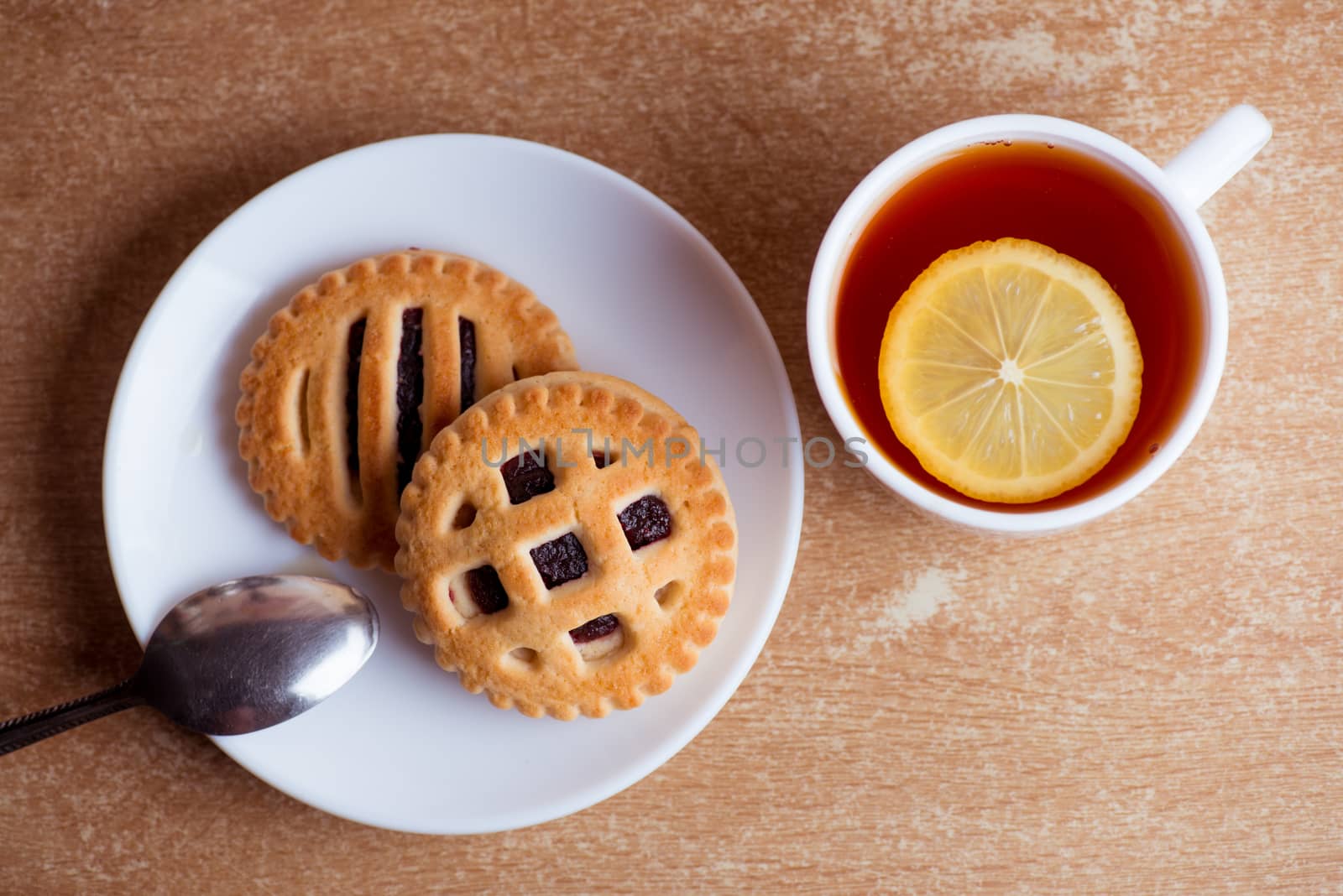 Cup of tea and lemon, cookies with jam in saucer on table top view.