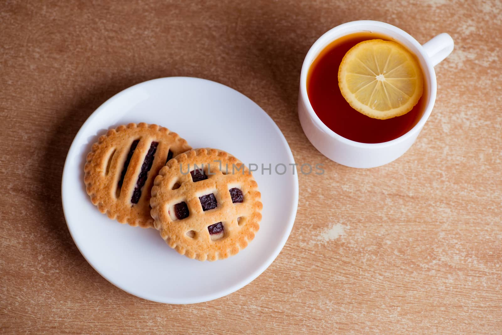 Cup of tea and lemon, cookies with jam in saucer on table top view.