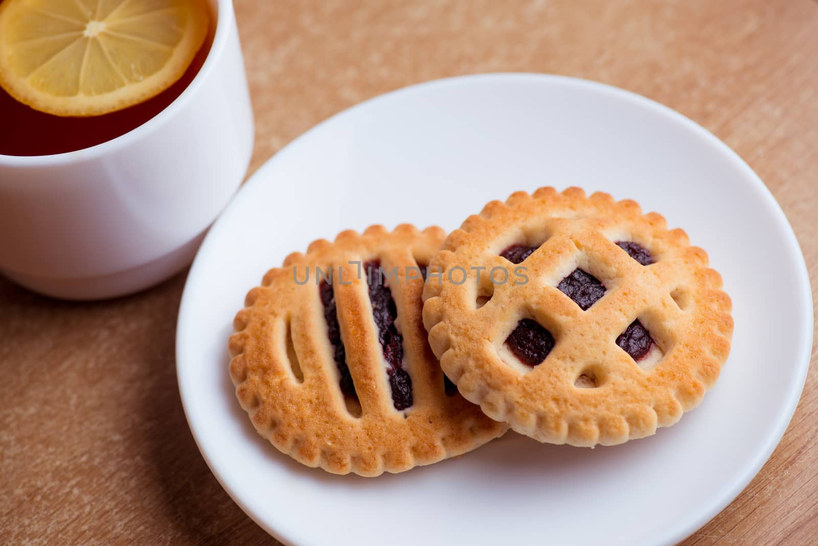 Cup of tea and lemon, cookies with jam in saucer on table top view.