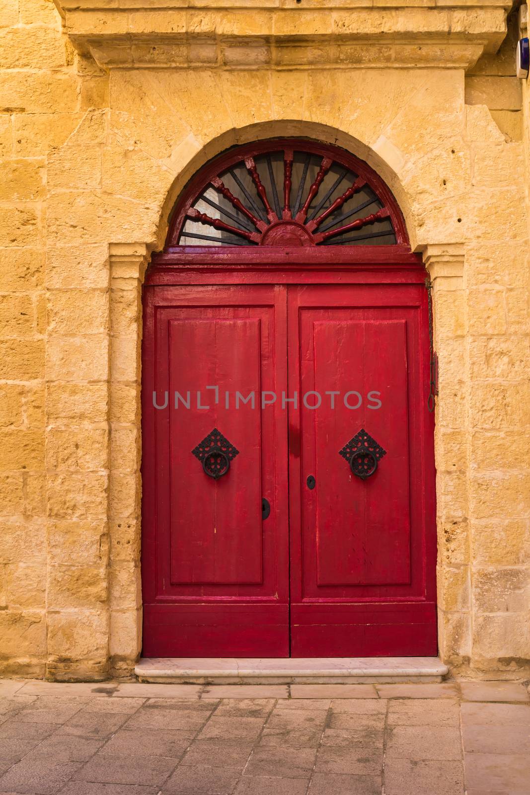 Former capital of island Malta, fusion of arabian and baroque architecture. Wall of a house made of stones with a ornate red door.