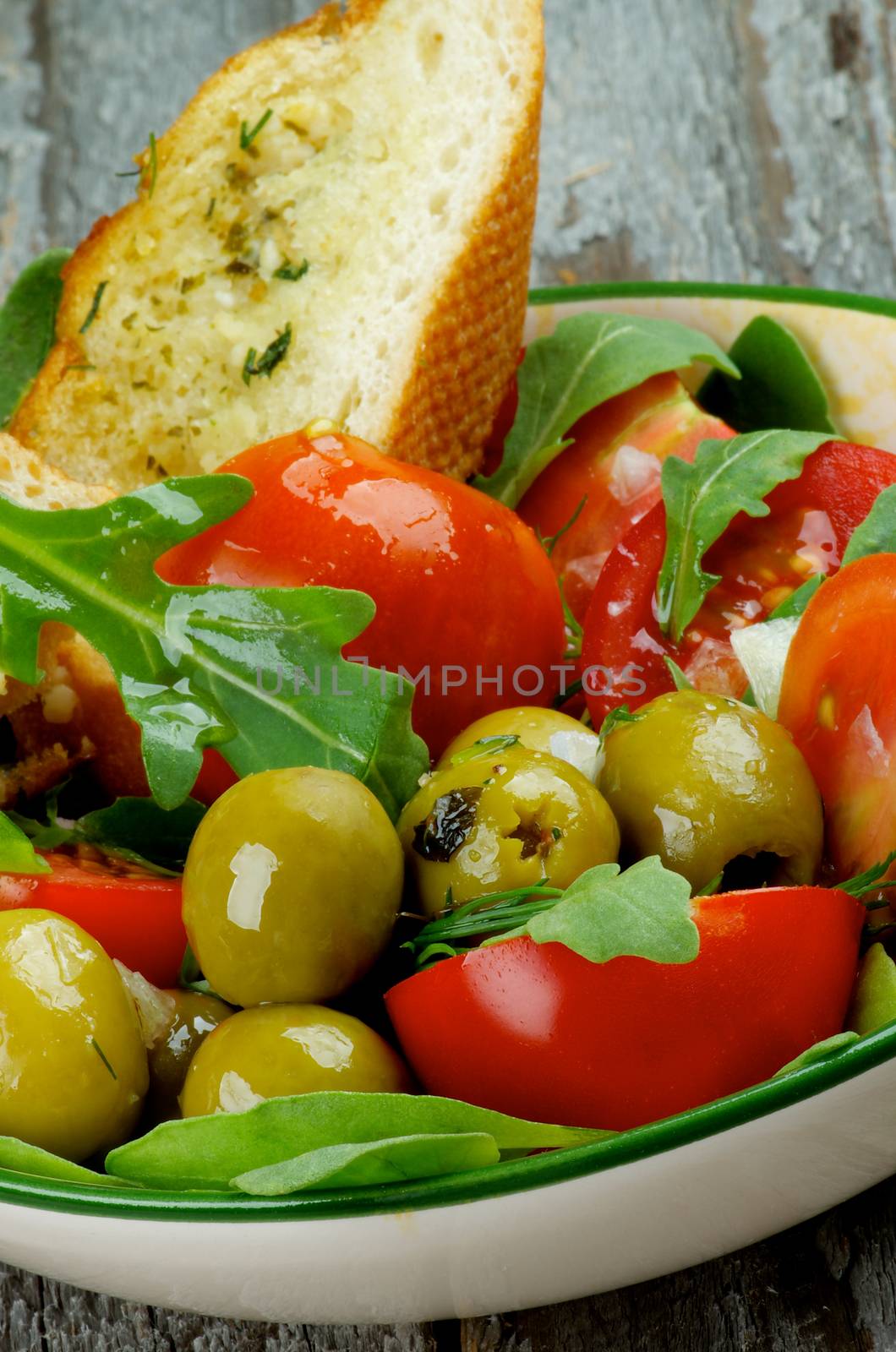 Fresh Tasty Tomatoes Salad with Arugula, Olives and Greens with Garlic Bread in Bowl closeup on Rustic Wooden background