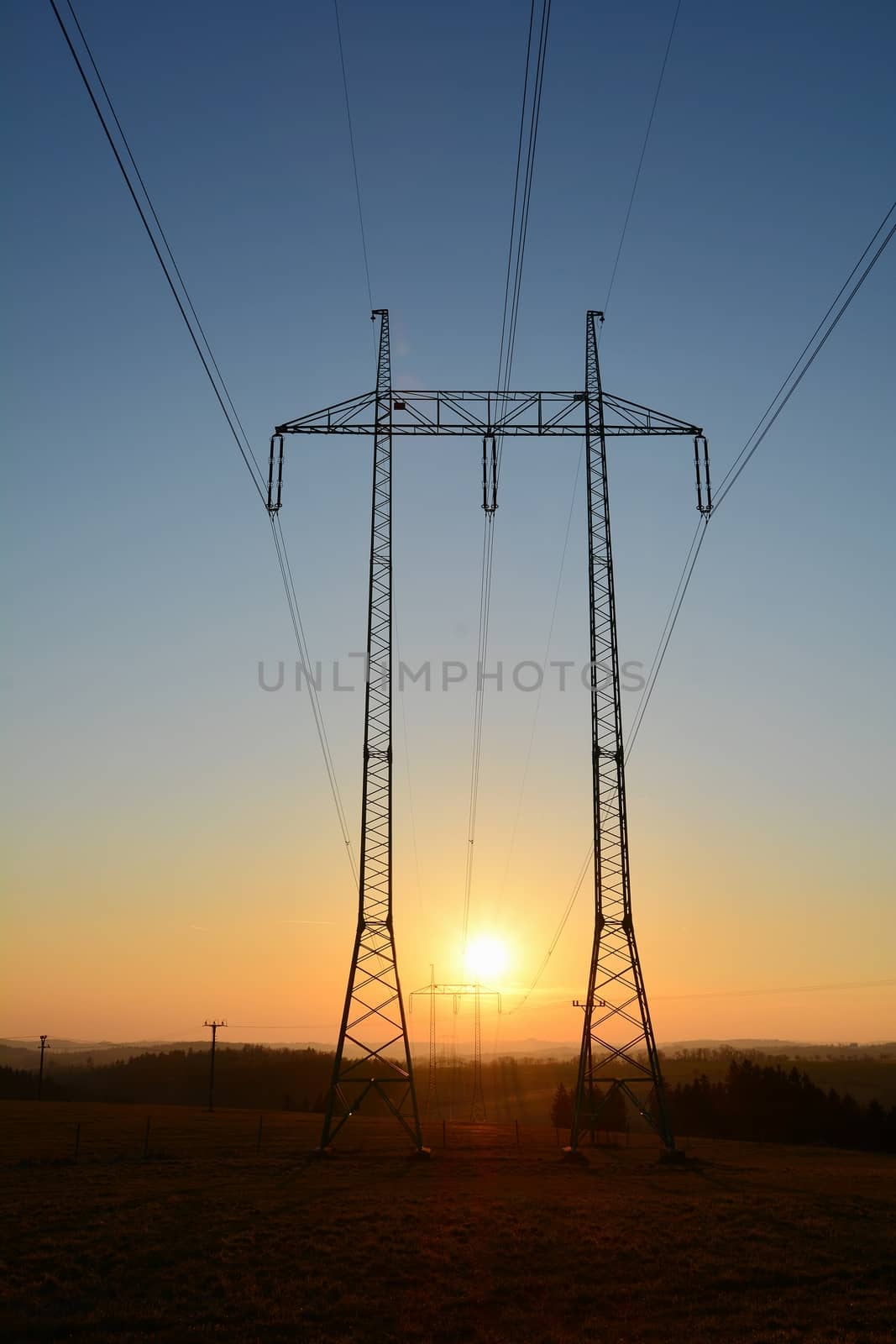 High voltage power lines with big and tall pylons during sunset. The sun sets between two pylons.