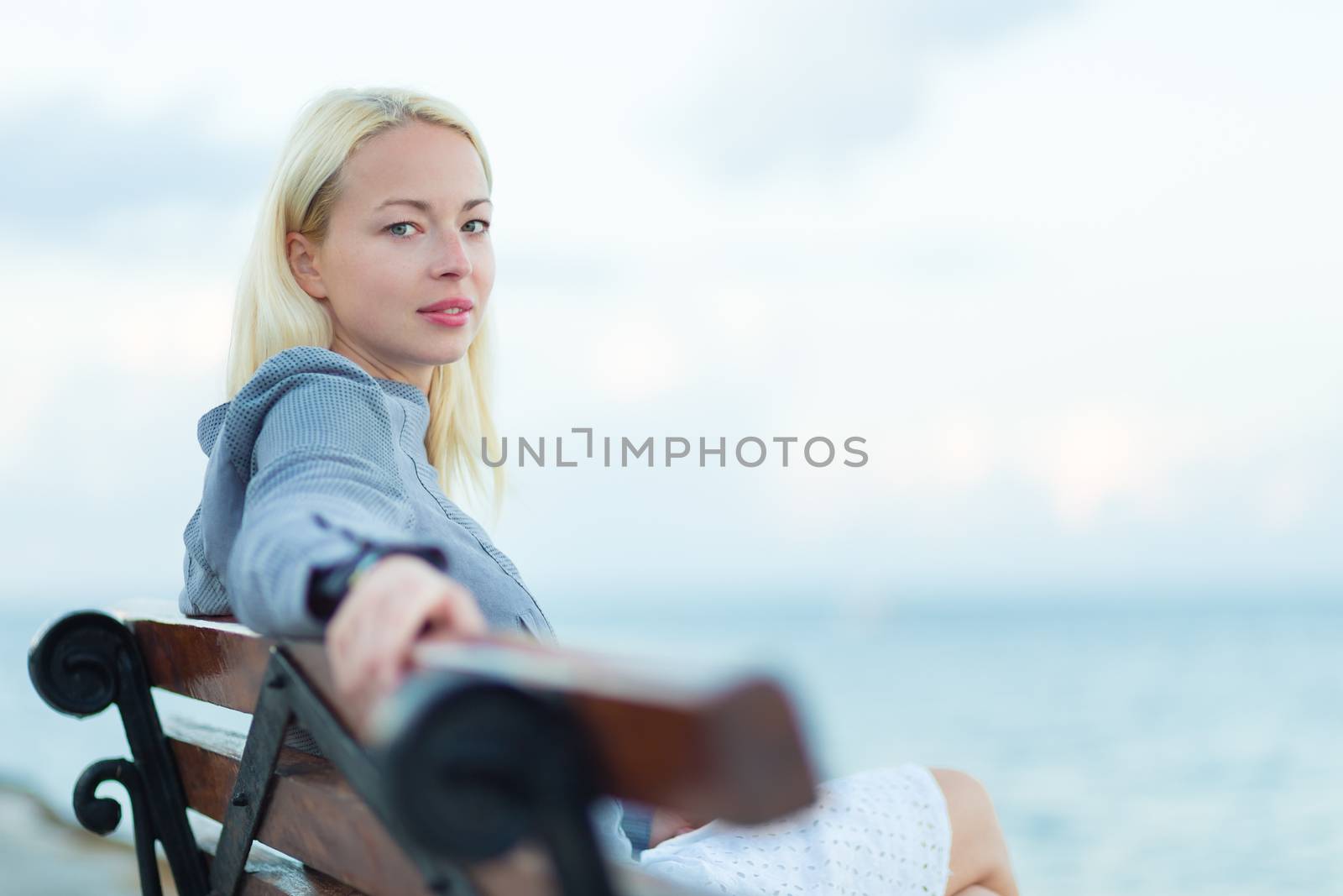 Beautiful woman sitting on a vintage wooden bench, relaxing on fresh breeze by the sea, looking at camera.