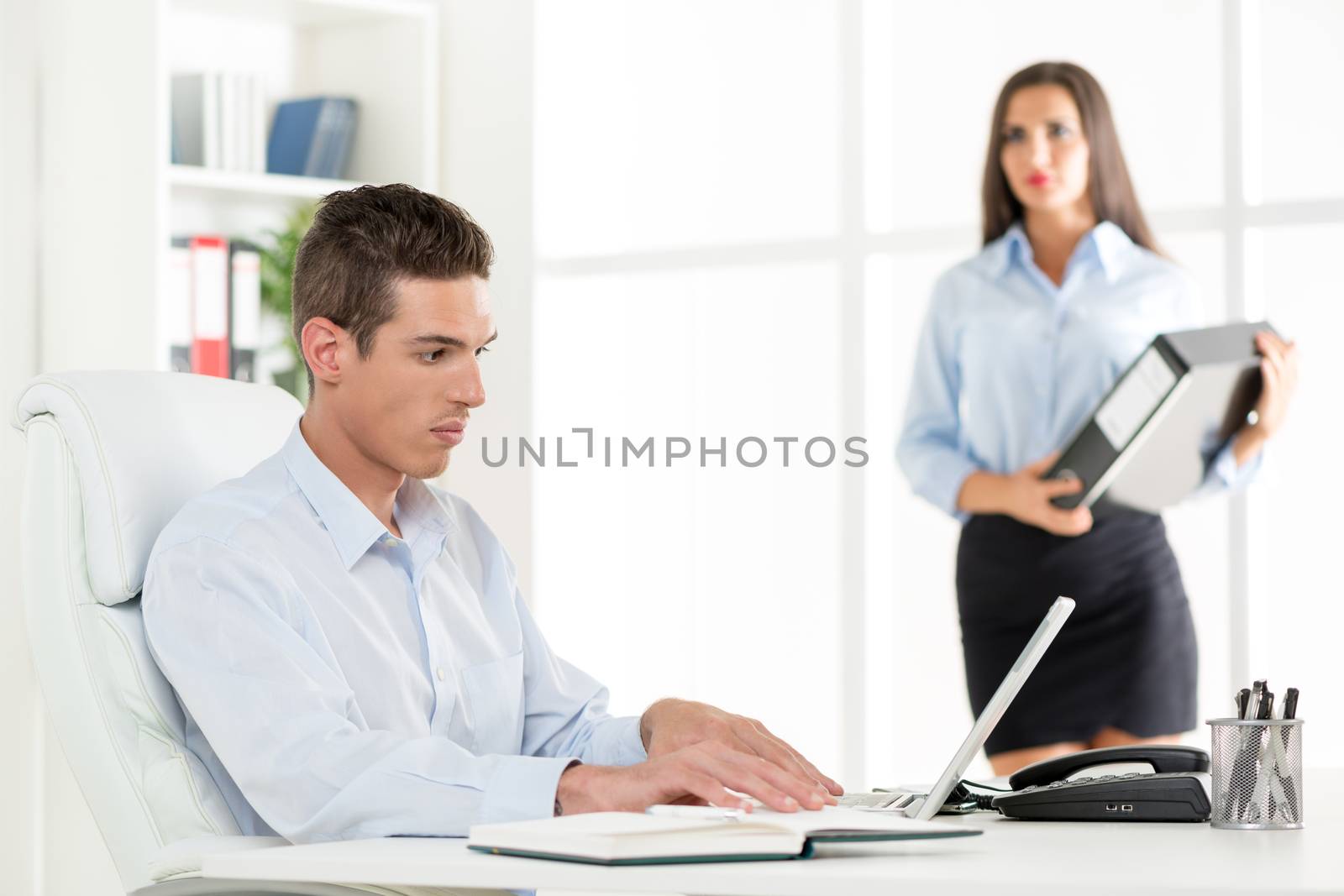 Beautiful young Businessman working on laptop in the office. Businesswoman standing with document in background. Selective focus.