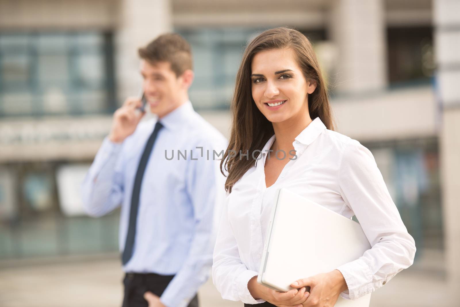 Young business couple standing in front of the building. Business woman carrying laptop while her partner use a mobile phone for business conversation.