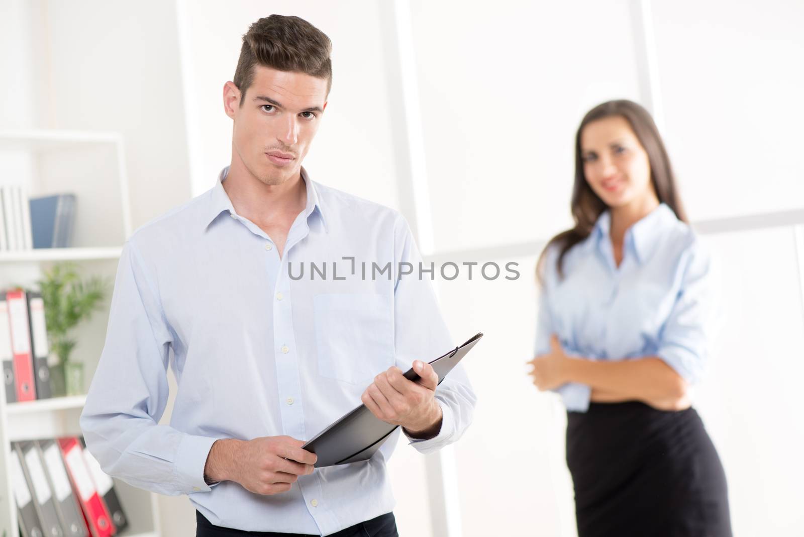 Beautiful young businessman standing with document in the office. Businesswoman standing in background. Selective focus.