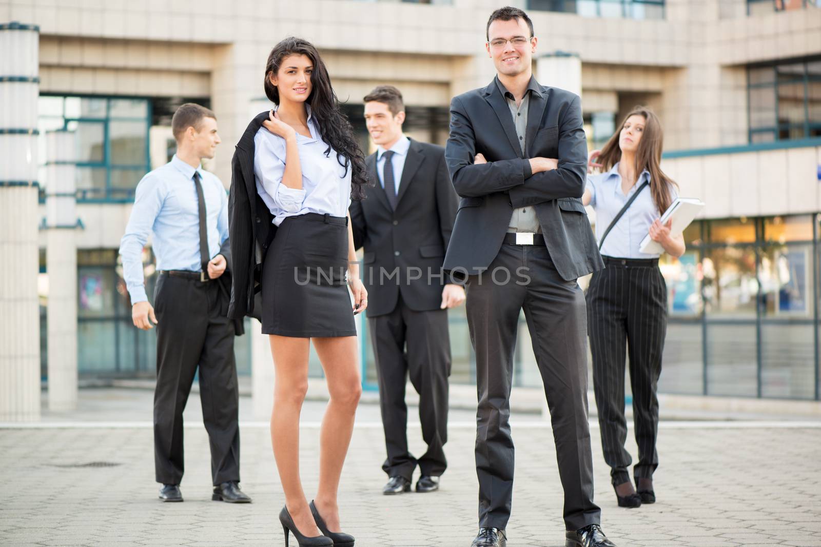 Young businesswoman and businessman with his team in the background standing in front of office building, looking at camera.