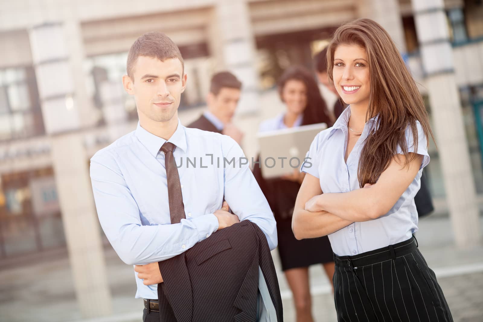 Young successful business couple with their business team in the background, standing in front of office buildings and with a smile looking at the camera.