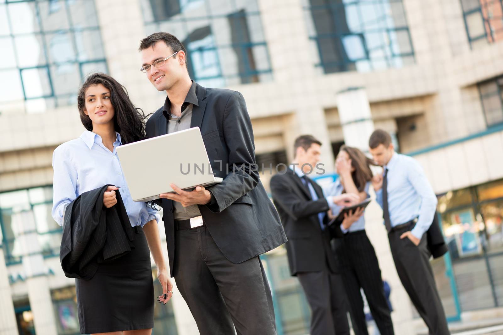 Smiling young businessman with a laptop and businesswoman standing in front of office building separated from the rest of the business team.