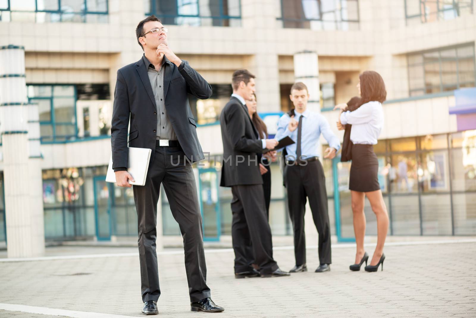 Young businessman standing in front of office building separated from the rest of the business team holding a closed laptop and happily looking into the distance, thinking about business success.