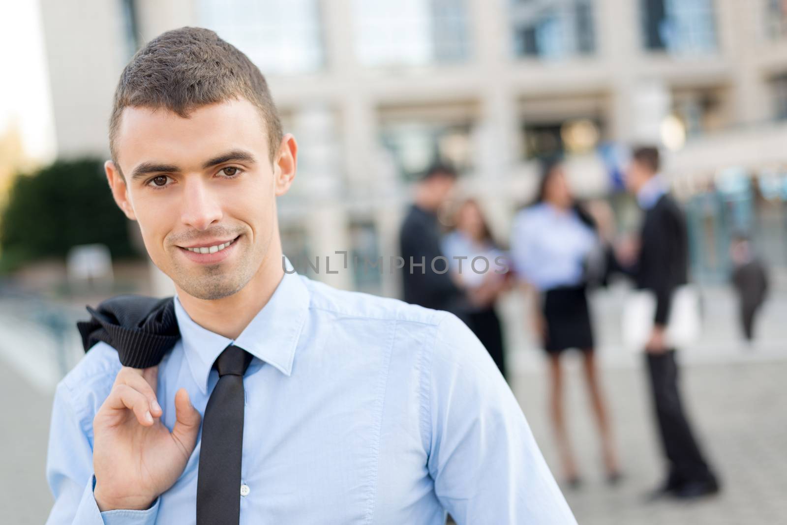 Portrait of a young businessman standing and holding coat draped over his shoulder looking into the camera with a smile. In the background is his business team and business building.