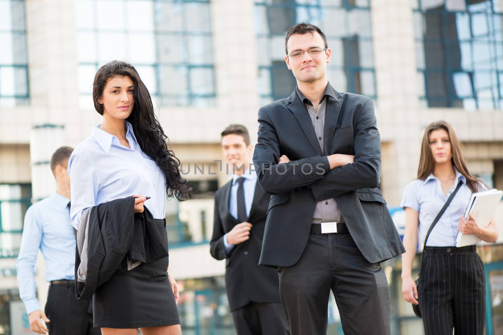 Young businesswoman and businessman with his team in the background standing in front of office building, looking at camera.