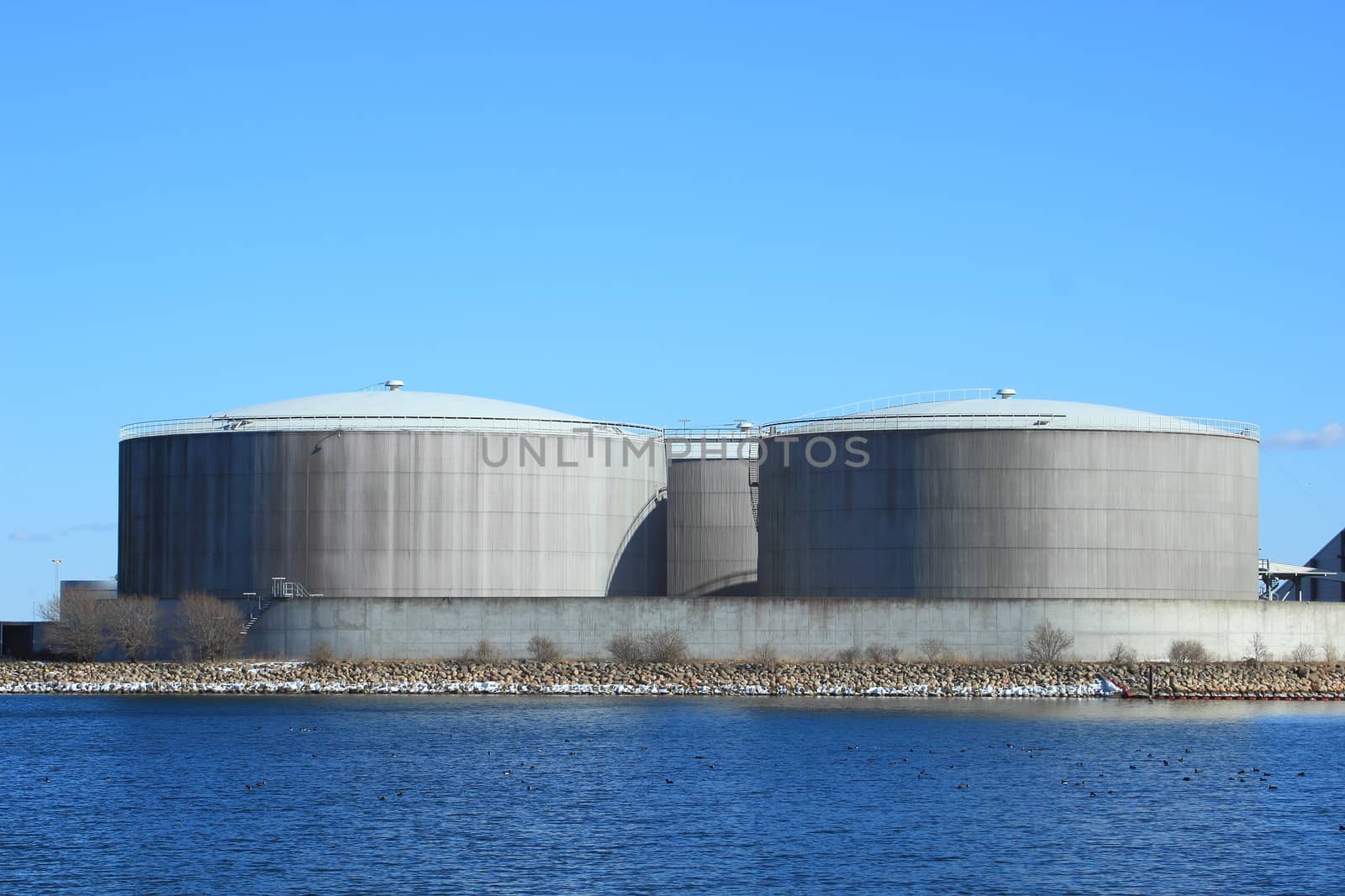 Gas containers at energy plant with lake and blue sky by HoleInTheBox