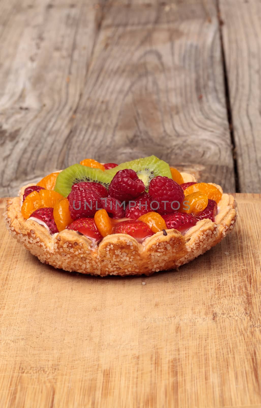 Fruit tart with a yellow custard, ladyfinger cookie shell crust, raspberries, strawberries, kiwi fruit and mandarin oranges on a cutting board on a rustic farm picnic table in summer.