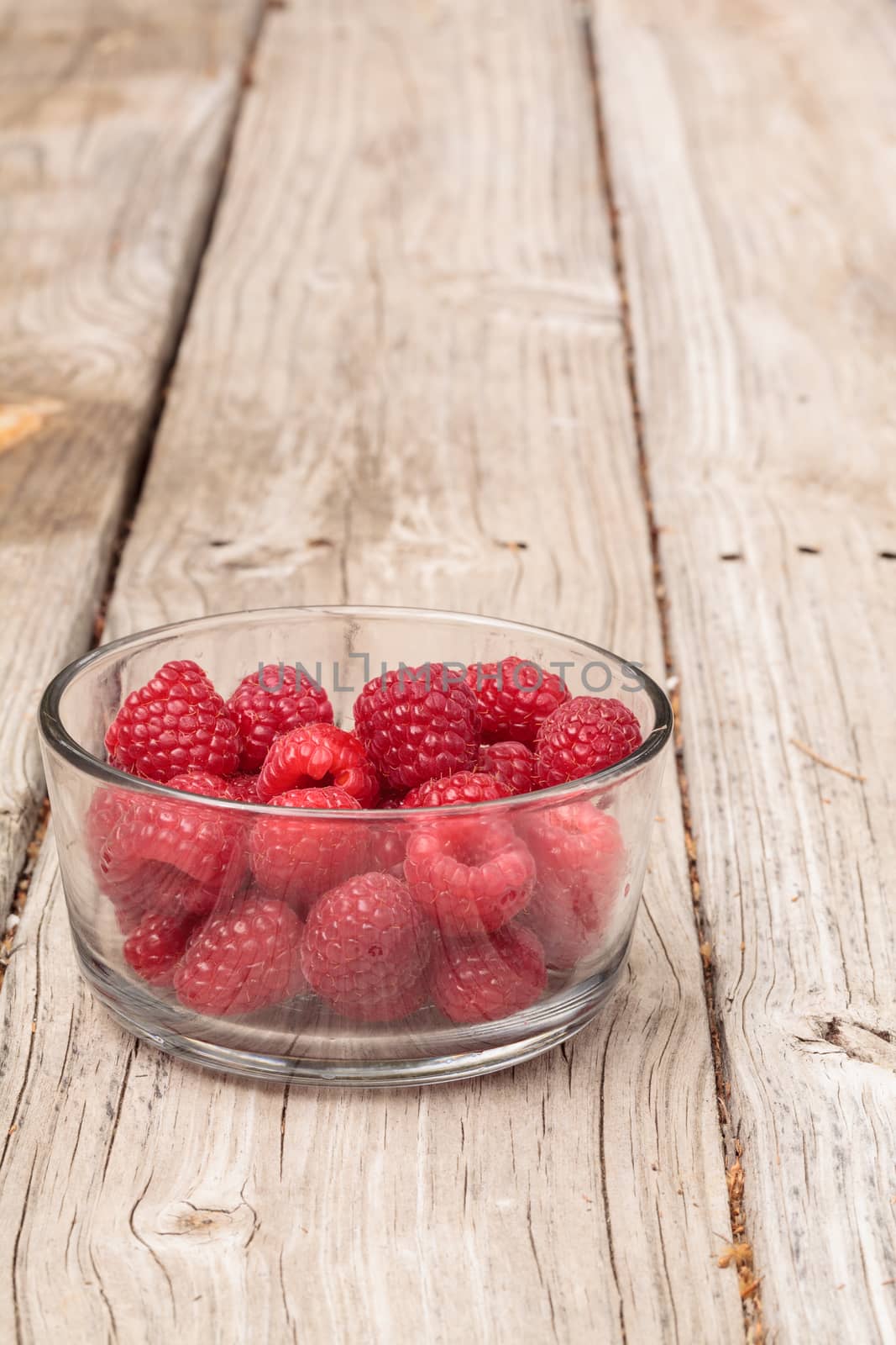 Clear glass bowl of ripe raspberries on a rustic farm picnic table in summer.