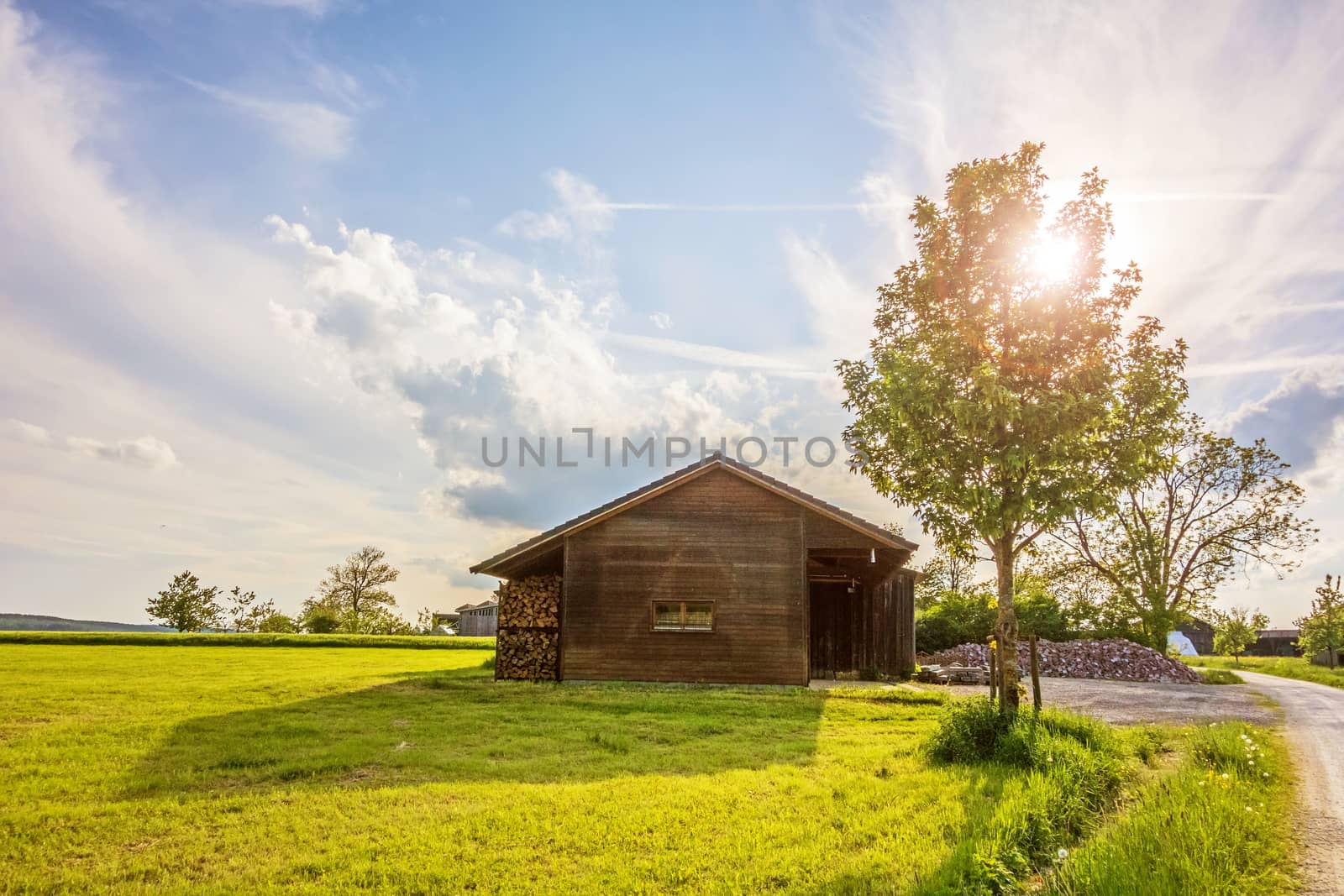 Old barn near farm at sunset - sun behind a tree, blue sky with clouds, meadow in front