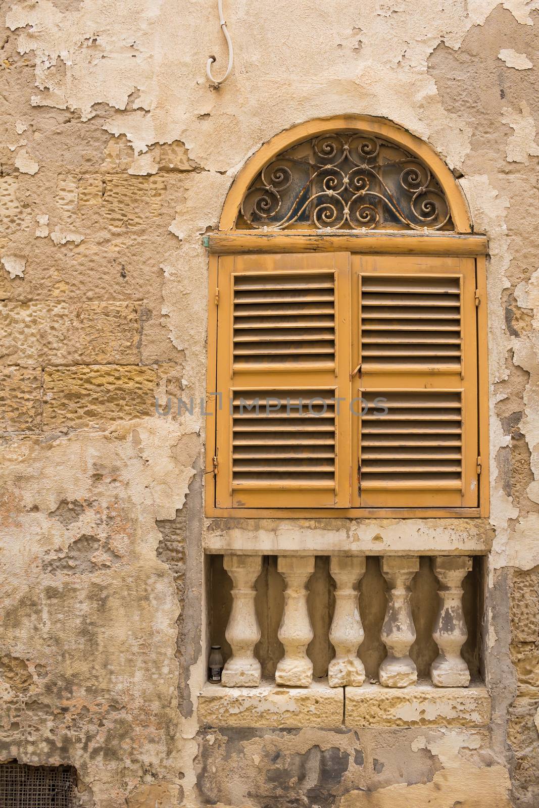 Facade of an old stone house. Window with an arch and yellow closed shutter. Decorative columns under the window. Senglea, island Malta.
