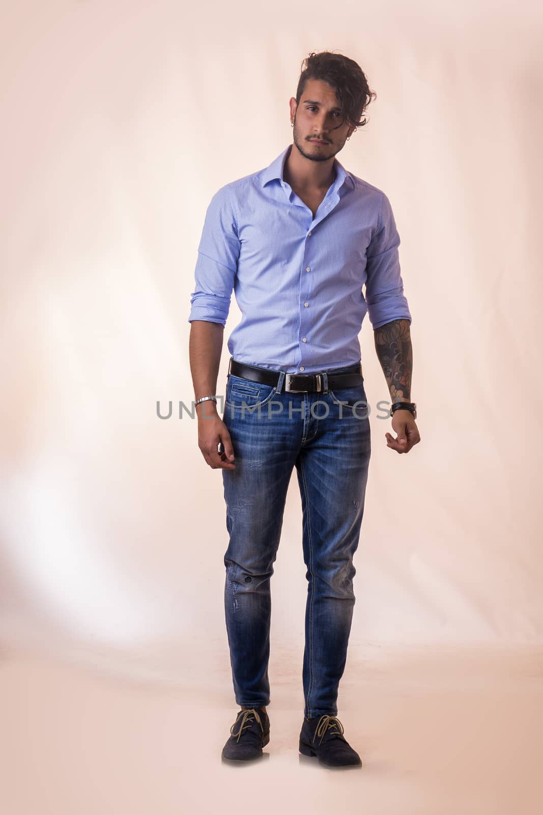 Portrait of brunette young man in light blue shirt and jeans, standing in studio shot against light background. Full length photo