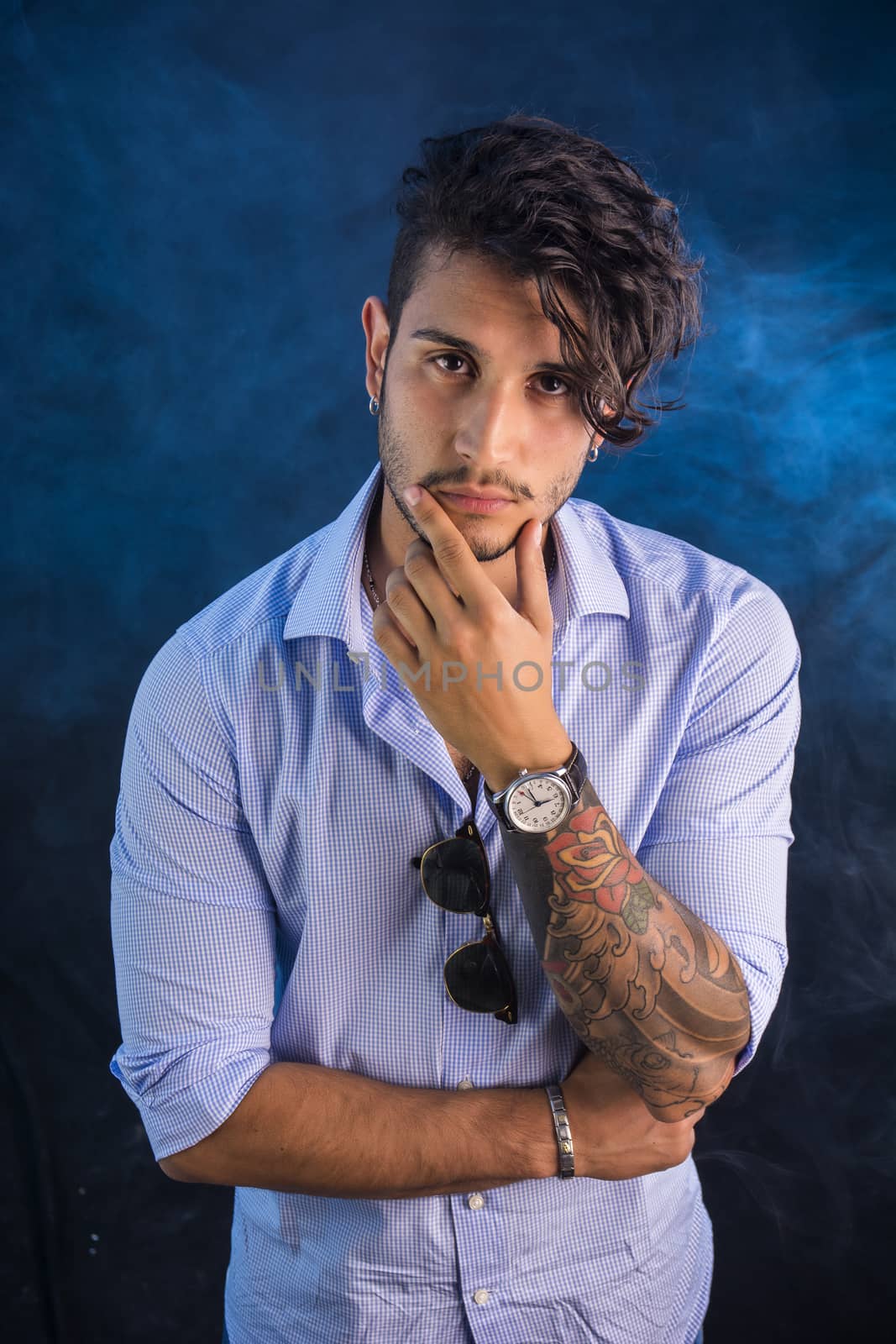 Portrait of brunette young man in light blue shirt and jeans, standing in studio shot against dark smoky background