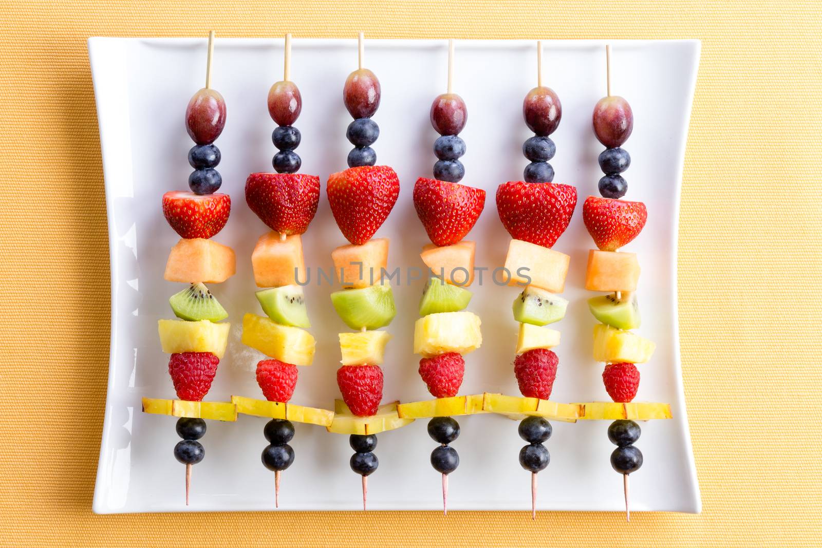 Healthy colorful fresh shish kebab fruit treats made from seasonal summer tropical fruit arranged neatly in a row on a modern white rectangular platter on a yellow textured table viewed from above