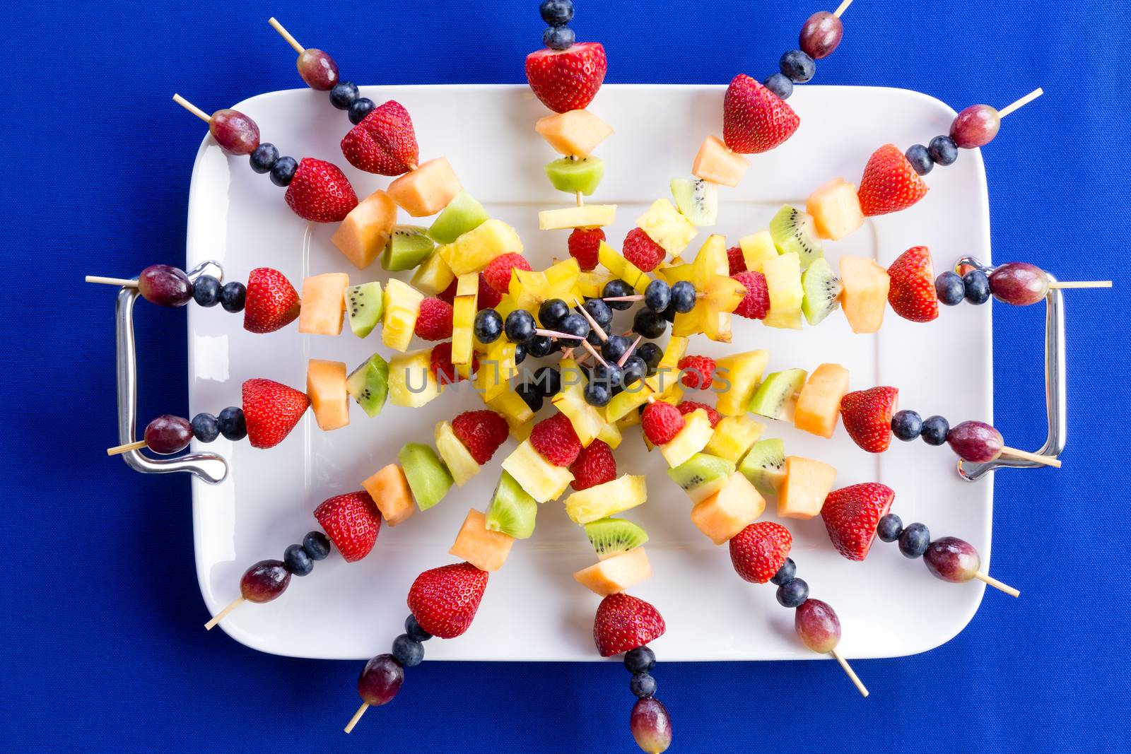 Colorful healthy fruit kebabs made with exotic and tropical fruit displayed in a radiating circle on a tray over a dark blue background, overhead view